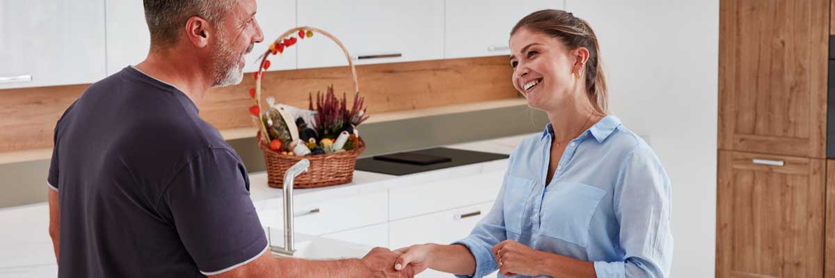 Reparatur- und Kundendienst Mann und Frau in der Küche, Hände schüttelnd, freundliches Gespräch, Küchenhintergrund mit Korb und Küchenmöbeln.